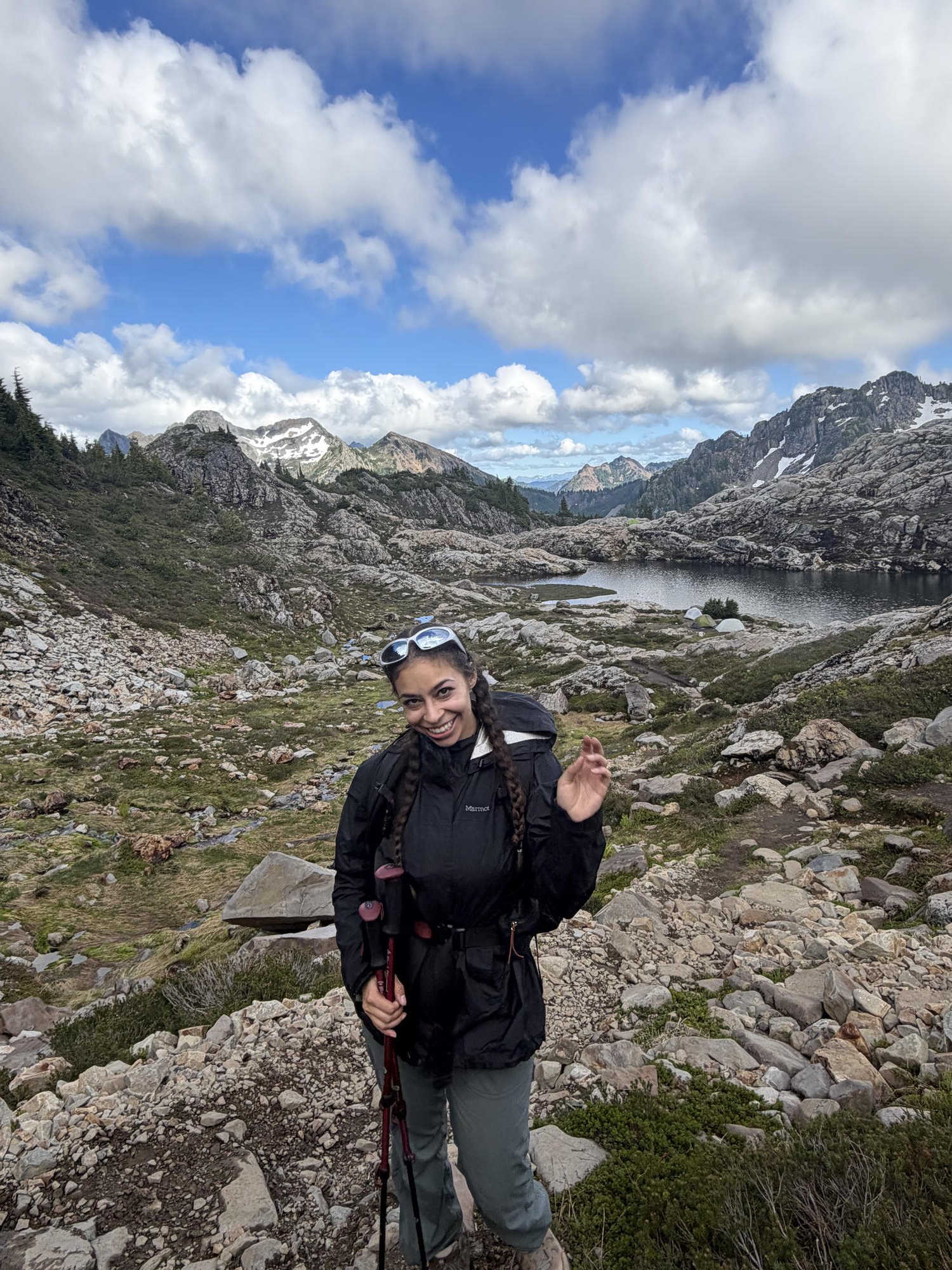 On the rocky alpine trail above the lake, trekking poles in hand, with mountains behind
