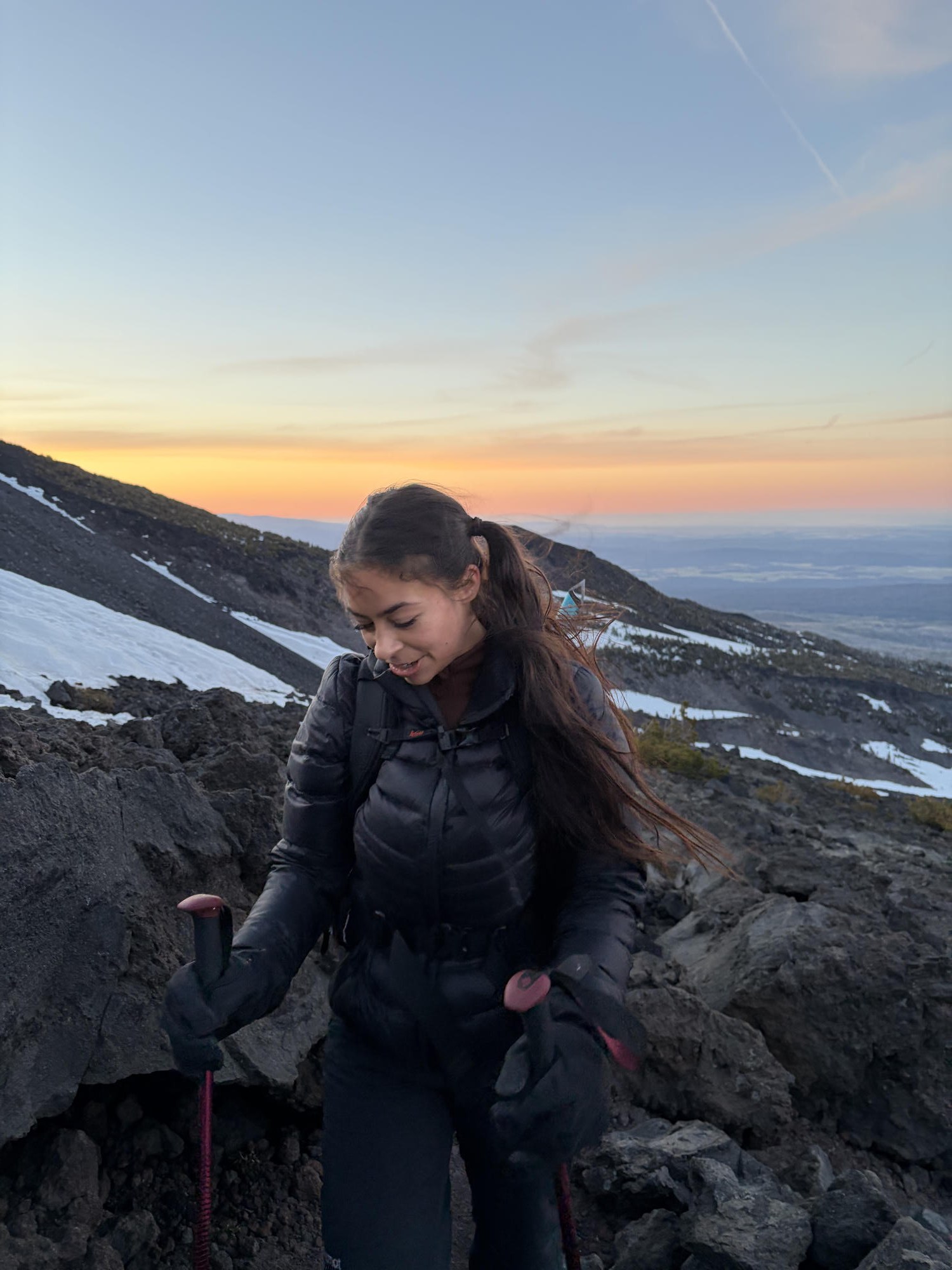 Smiling on the rocky slope with trekking poles as the sun rises in a glowing orange band behind